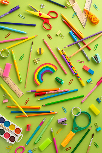 Emphasize the back-to-school theme: Vertical top view of scattered color pencils, watercolor paints, plasticine, scissors, ruler, clips, magnifying glass, erasers and more on pastel green background