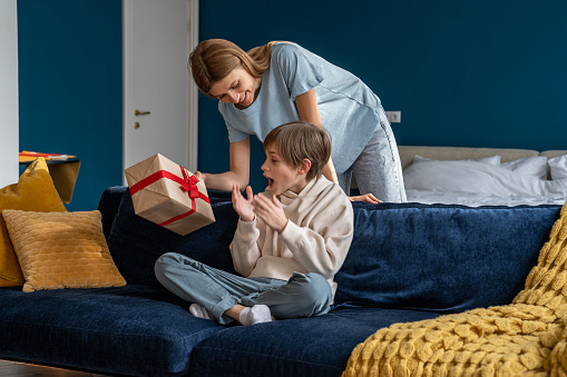 Young woman loving mother surprising son with birthday gift at home, mom giving wrapped box to excited little boy, congratulating child. Overjoyed happy kid sitting on sofa getting present from mum