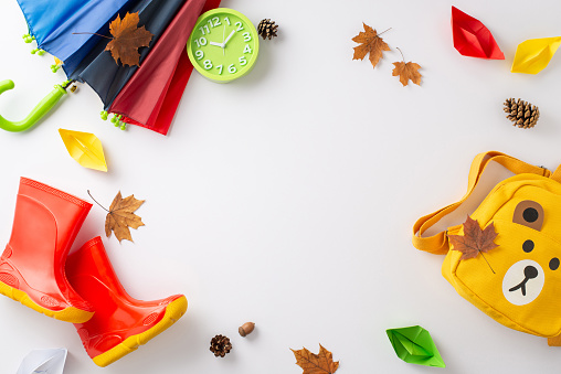 Delightful rainy day scene in autumn for children's joy. Capture the moment with a top view image of a colorful umbrella, gumboots and yellow backpack on a white backdrop with copy-space