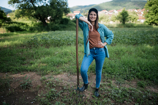 Portrait of a happy woman farmer standing with pitchfork in the farm. Female worker holding a gardening tool looking at camera and smiling at potato crop field.