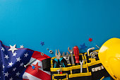 Labor Day tribute concept. Top view of USA flag, glitter stars, construction tools, screwdriver, hard hat, tool belt, wrench, needle nose pliers, nails, nuts, screws, hex keys set on blue backdrop