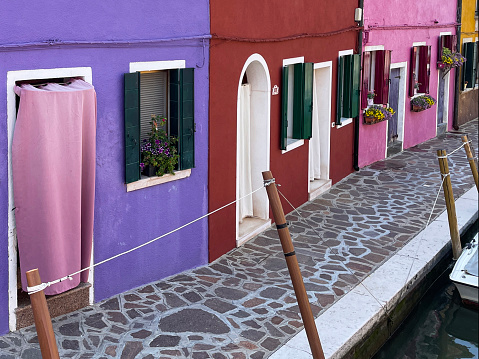 Stock photo showing view of an alley lined with colourful buildings beside a canal and moored boats in Burano, Venice, Italy. Famous for lace making, Burano is an island in the Venetian Lagoon.