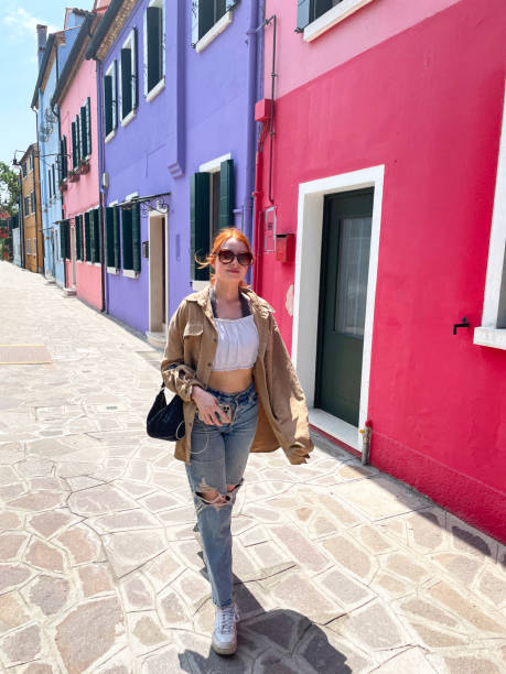 Image of attractive, redheaded woman wearing sunglasses, walking through alleyways of Burano, Venice, Italy, looking at colourful buildings and architecture, focus on foreground Stock photo showing close-up view of beautiful, red haired, female tourist wearing white crop top, fleece jacket and ripped jeans whilst walking through Burano alleyways. Famous for lace making, Burano is an island in the Venetian Lagoon. crop top view stock pictures, royalty-free photos & images