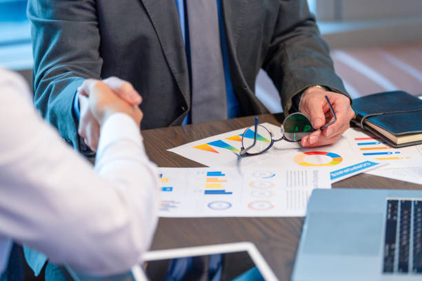 istock Businessmen shaking hands. Both are dressed in business clothing and there is paperwork with finance graphs and charts. 1568200367