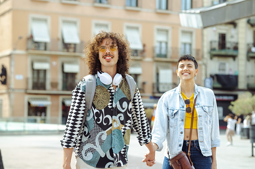 A young Spanish-Portuguese man and a mid-adult Spanish woman are holding hands and cheerfully walking down the streets of Barcelona.