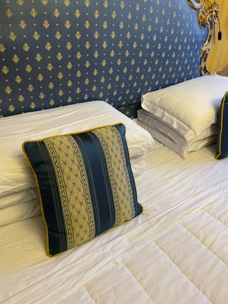 istock Full frame image of hotel room double bed with stack of white hotel pillows leaning on ornate framed, patterned, padded headboard, white duvet cover, blue and gold striped cushions, focus on foreground 1568251830