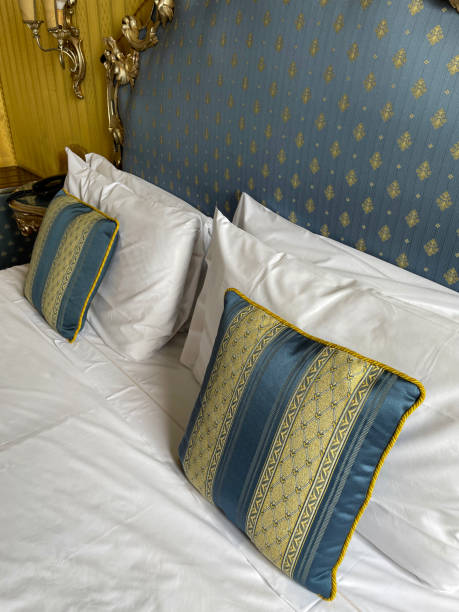 istock Full frame image of hotel room double bed with stack of white hotel pillows leaning on ornate framed, patterned, padded headboard, white duvet cover, blue and gold striped cushions, wall mounted electric lamp, night stand, focus on foreground 1568252377