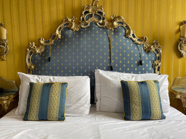 Stock photo showing close-up view of gold and blue striped cushions leaning against a pile of white hotel pillows resting on ornate framed, patterned, padded headboard on hotel room duvet bedding. imagen en primer plano de la cama doble de la habitación del hotel con una pila de almohadas blancas de hotel apoyadas en una cabecera ornamentada enmarcada, estampada y acolchada, funda nórdica blanca, cojines a rayas azules y doradas, lámparas eléctr