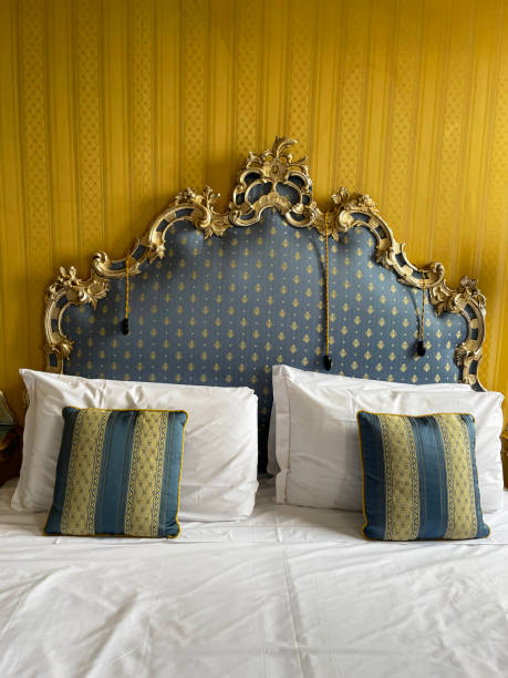 istock Image of hotel room double bed with stack of white hotel pillows leaning on ornate framed, patterned, padded headboard, white duvet cover, blue and gold striped cushions, focus on foreground 1568252897
