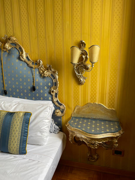 istock Image of hotel room double bed with stack of white hotel pillows leaning on ornate framed, patterned, padded headboard, white duvet cover, blue and gold striped cushion, wall mounted electric lamp, night stand, focus on foreground 1568252900