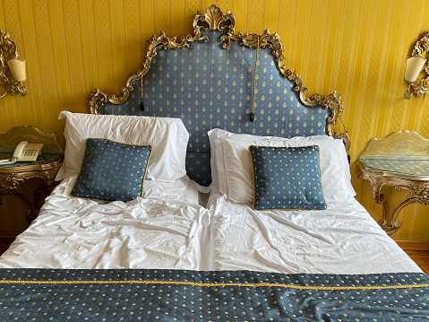 Stock photo showing close-up view of gold and blue striped cushions leaning against a pile of white hotel pillows resting on ornate framed, patterned, padded headboard on hotel room duvet bedding.