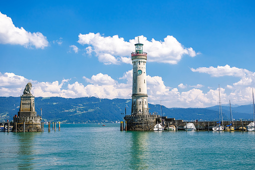 Lindau, Bavaria, Germany. Old lighthouse with clock in bay. Antique bavarian town at Lake Constance Bodensee. Monument with statue of lion at entrance to port, yachts by piers. Summer landscape.