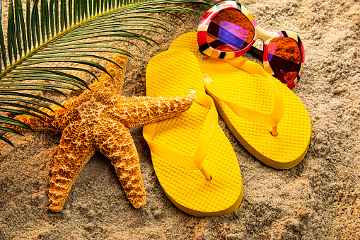 This is a photograph of yellow sandals, sunglasses and a palm branch on a sandy beach