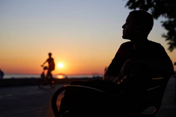 silhouette portrait of a young man sitting at a park bench near seaside and waiting