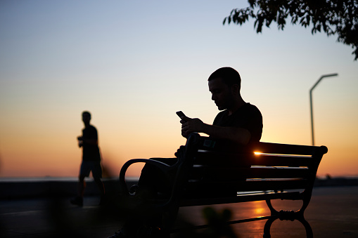 silhouette portrait of a young man sitting at a park bench near seaside and waiting