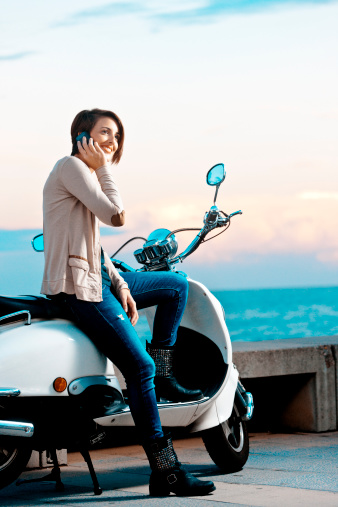 A young woman leaning against her moped bike, talking on a mobile phone. Sea in the background, Barcelona, Spain