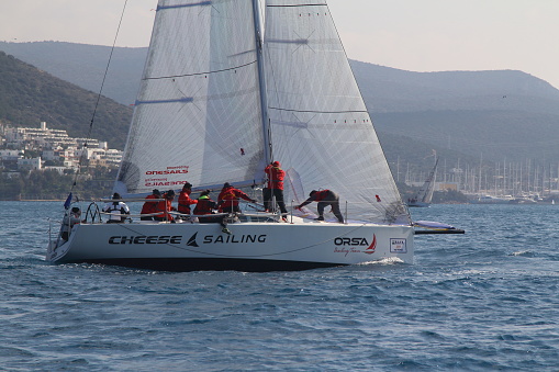 Bodrum,Mugla, Turkey. February 09,2019 sailor team driving sail boat in motion, sailboat wheeling with water splashes, mountains and seascape on background. Sailboats sail in windy weather in the blue waters of the Aegean Sea, on the shores of the famous holiday destination Bodrum