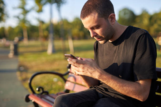 istock young man sitting on a park bench and messaging 1568353009