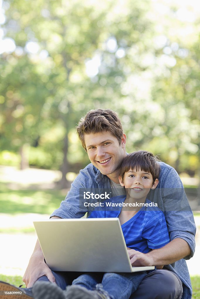 Sorridente menino e seu pai sentados tanto usando laptop - Foto de stock de 30 Anos royalty-free