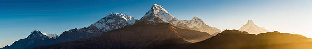 Golden rays of dawn light shining through the early morning mist and illuminating the iconic snow capped peaks of the Annapurna range and Machapuchare from the famous Poon Hill viewpoint under clear blue Himalayan skies, Nepal. ProPhoto RGB profile for maximum color fidelity and gamut.