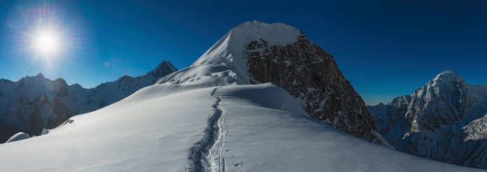 Mountaineers moving roped together along a snowy col towards the final summit headwall of Tharpu Chuli (5663m) beneath bright sunlight flaring in blue high altitude skies and surrounded by the iconic peaks and pinnacles of the Annapurna Conservation Area deep in the Himalayan mountain wilderness of Nepal. ProPhoto RGB profile for maximum color fidelity and gamut.