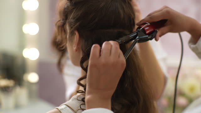 Closeup of process of curling long hair with hair tongs in modern beauty studio in front of mirror. Close up shot of hairstylist curling hair using hair iron.