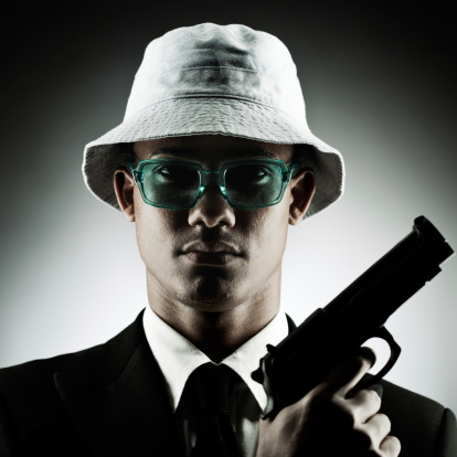 One young man holding a handgun. More files of this model and theme on port. Square composition. He is formally dressed and wearing a funny hat and glasses in a dark background.