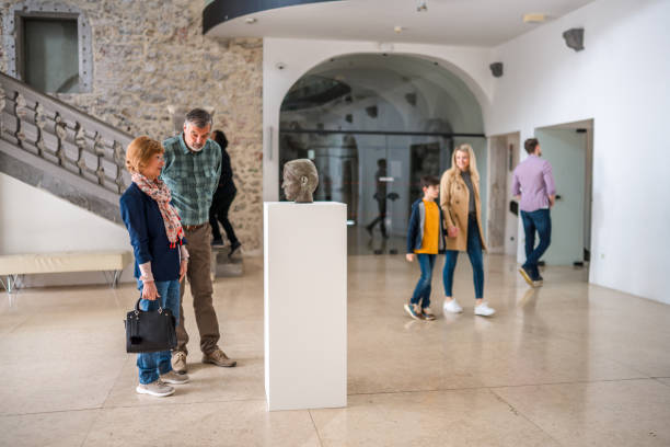An elderly couple looking at a sculpture on display at a big, open exhibition space with various people passing, bright light, full length shot.