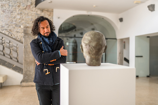 Mid adult, black-haired, curly man standing in front of a bronze object on a white pedestal, looking at it attentively. 3/4 length shot, arms crossed over his chest.