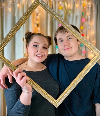 Cute teenage couple at a party with a homemade Photo Booth, holding up a frame and posing arm and arm inside and having fun. Lighted curtain backdrop.