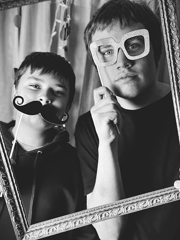 Two teen boys at a Homemade Photo Booth Party. The boys are holding Photo Booth props and having fun. Lighted curtains in the background. Photo is in black and white.