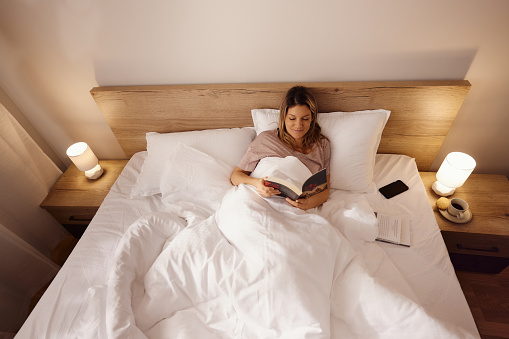 High angle view of young smiling woman reading a book during morning in a bed.