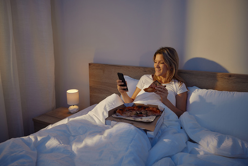 Young happy woman using mobile phone while eating fresh pizza in a bed. Copy space.