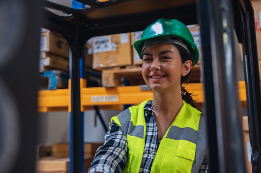 Experience the dynamic atmosphere of a distribution warehouse as a young, happy female forklift driver takes charge of operations. This captivating image showcases her confidence and skill as she poses and smiles while driving the worklift. Embrace the industrial and industrial concept, where efficiency and productivity are paramount. Witness the seamless coordination between the driver and the forklift, reflecting the dedication and expertise necessary in the warehouse environment. This image captures the spirit of success and highlights the pivotal role of skilled workers in ensuring smooth operations.