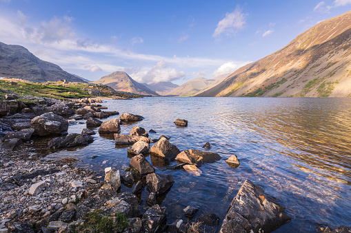 A photograph taken at Wastwater in the English Lake District on a fresh summer morning. The sun can be seen rising through the peaks of the mountains, creating a starburst sun ray effect.