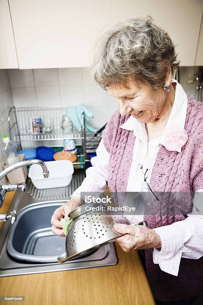 Anziana signora sorridente lavaggi un colapasta presso kitchen sink - Foto stock royalty-free di Lavare i piatti
