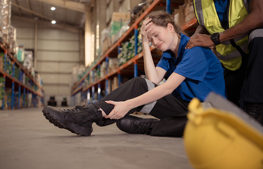 A warehouse worker consoles and helps a female worker who cries out in pain after a leg accident in a large warehouse