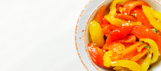 Sliced red and yellow roasted peppers with garlic and parsley in a white ceramic bowl. Concept Traditional Italian dish. Vegetarian and vegan food. Horizontal orientation. Top view. Copy space Banner