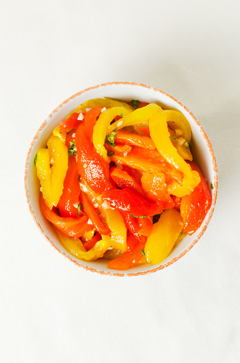 Salad with red and yellow roasted pepper slices, garlic and parsley in a white ceramic bowl. Concept of healthy eating. Traditional Italian dish. Vegetarian and vegan food. Vertical orientation.