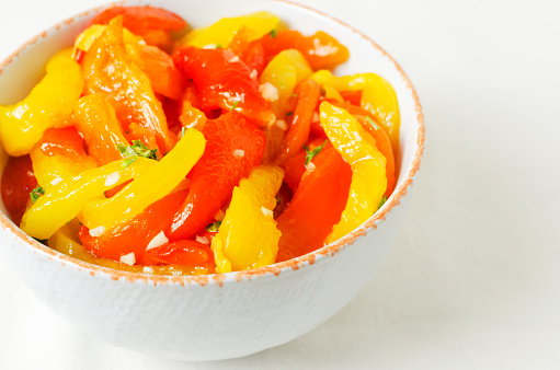 Salad with red and yellow roasted pepper slices, garlic and parsley in a white ceramic bowl. Concept of healthy eating. Traditional Italian dish. Vegetarian and vegan food. Horizontal orientation.