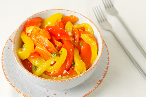 Salad with red and yellow roasted pepper slices, garlic and parsley in a white ceramic bowl. Concept of healthy eating. Traditional Italian dish. Vegetarian and vegan food. Horizontal orientation.