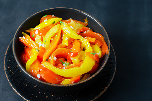 Salad with red and yellow roasted pepper slices, garlic and parsley in a black ceramic bowl on a black background. Concept of healthy eating. Traditional Italian dish. Vegetarian and vegan food.