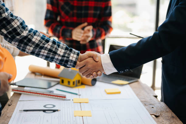 istock Construction team shake hands greeting start new project plan behind yellow helmet on desk in office center to consults about their building project. 1568985189