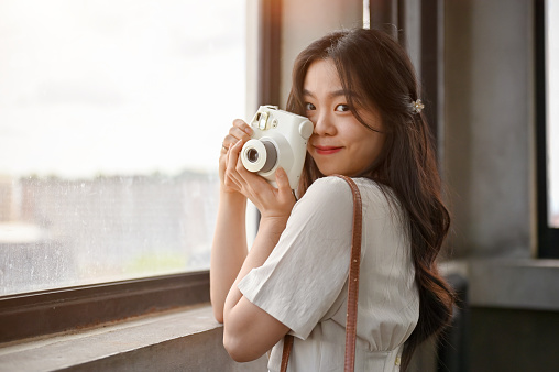 A portrait of a beautiful Asian female tourist in casual clothes standing by the window with her instant camera, looking at the city view from a city tower. travel, tourist attractions, lifestyle
