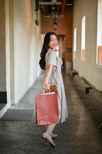 A portrait of an attractive and happy Asian female tourist carrying her shopping bags, walking inside the building, enjoying her weekend in the city. Lifestyle concept