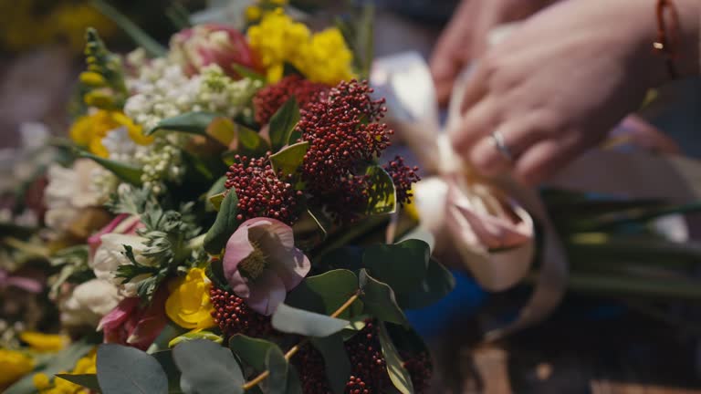 Close-up bouquet with blurred female hands tying pink bow on green stems at background. Closeup unrecognizable Caucasian young woman making gift in flower shop indoors.