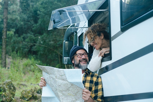 Happy mature couple enjoy vanlife camper vacation travel lifestyle talking and looking together a map guide to choose next holiday destination. Man outside and woman at the window. Motorhome vehicle