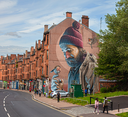 Glasgow, Scotland - 23 Junkly 2023: A mural by the Australian artist smug (Sam bates) on a gable end of a tenement block in Glasgow's High Street depicting a modern interpretation of the city's saint, St Mungo, saving a robin. Three people are having a close up view of the mural while other pedestrians are walking along the pavement.