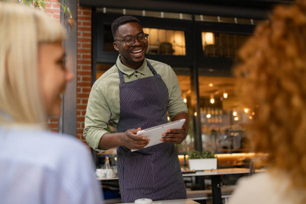 istock Happy black waiter taking orders from customers in cafeteria. 1569143162