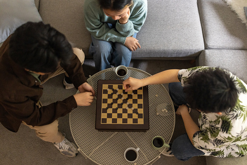 Two young Chinese men playing checkers in the living room of a house in the company of their female friend